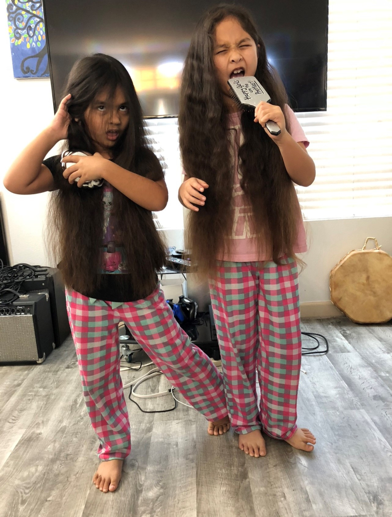 Two young girls with long brown hair wearing pink pajamas pretend to sing into hairbrushes at home