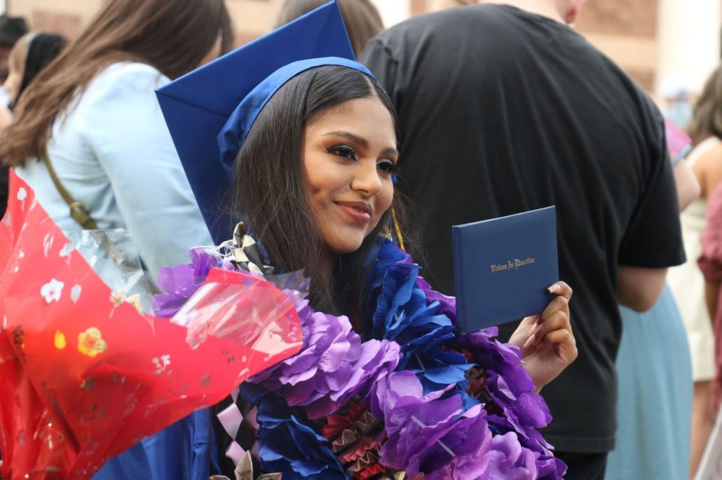 Dark haired female graduate poses with her diploma in flower leis and a cap and gown at graduation