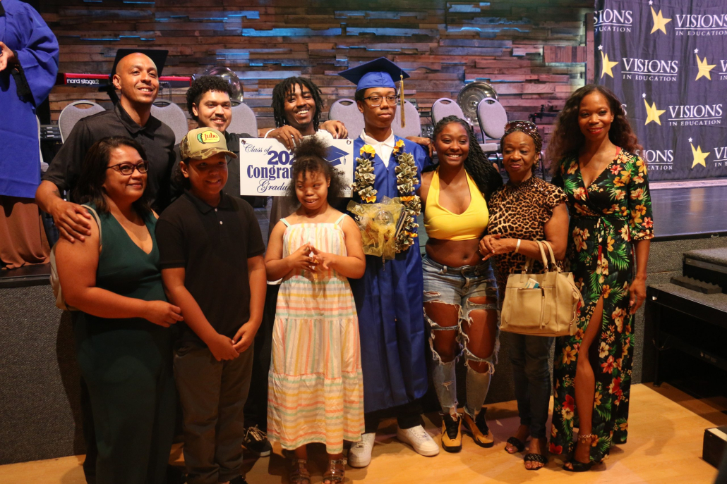 A graduate in a blue cap and gown stands and smiles for a photo with a large family.
