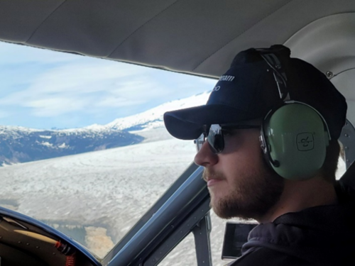 Hunter Sites web In a navy cap, black sunglasses and green over-the-ear headset, Hunter sits in the cockpit of a plane flying over clouds and looking out over the horizon.