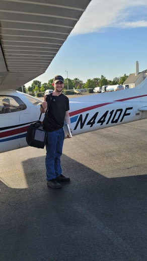 Sites 3 In a cap, glasses, a black shirt and jeans, Hunter stands next to a plane on the runway with his gear and a book in his hand, giving a thumbs up to the camera.