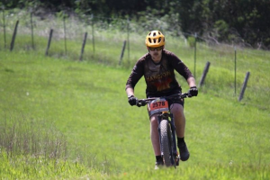 In a yellow bike helmet, Hunter rides his cross-country mountain bike over a big, green meadow with a brown fence and foliage in the background.