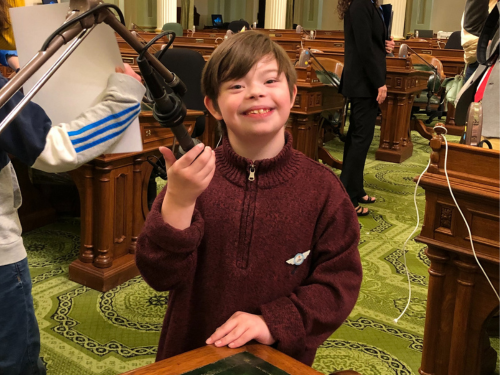Dominic www In a maroon-colored sweater, Dominic smiles for the camera as he holds a podium microphone at the California State Capitol.