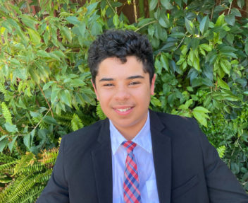 In a black suit, light purple shirt and red and blue tie, Bassam poses for a photo in front of a bright green bush.