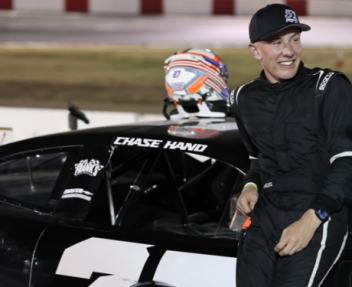 Chase Hand, wearing a black had and all-black racing suit, leans against a black race car with his name and number 27 on it.