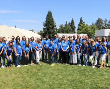 Large group of Visions staff in matching light blue t-shirts stand together on a lawn and smile for a photo.