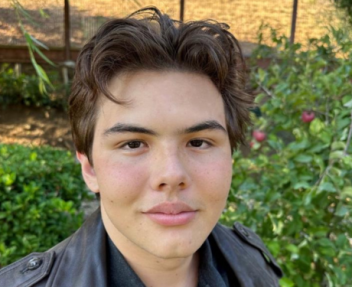 Selfie of a teenage boy with brown hair wearing a leather jacket in front of green bushes.