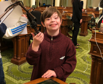 In a maroon-colored sweater, Dominic smiles for the camera as he holds a podium microphone at the California State Capitol.