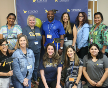 A group of male and female Visions employees smile in front of a blue, yellow and white backdrop that says Visions In Education.