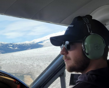 In a navy cap, black sunglasses and green over-the-ear headset, Hunter sits in the cockpit of a plane flying over clouds and looking out over the horizon.