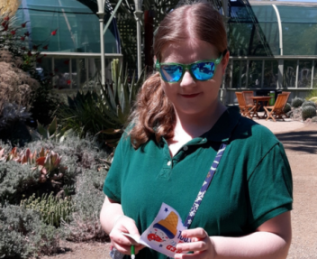 Teenage girl with a side ponytail wearing blue reflective sunglasses and a purse diagonally across her body looks down at a pamphlet in her hands.