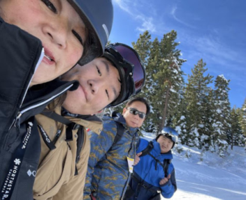 A family of four smile for a photo in snow gear as they ride on the chair lift of a ski resort.