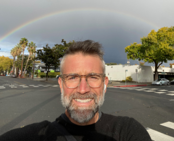 Mitch takes a selfie wearing a black t-shirt and Apple Airpods standing underneath a rainbow.