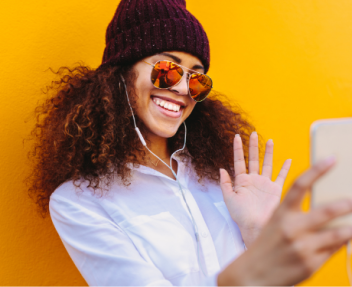 Female high school students waves into iphone taking a selfie wearing beanie and sunglasses against yellow background