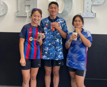 Three smiling home school students in soccer jerseys pose with Boba drinks in their hands in front of a wall at their family-owned cookie business