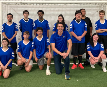 Visions students and soccer club members in blue jerseys gather in front of the soccer net on the turf for a group photo.