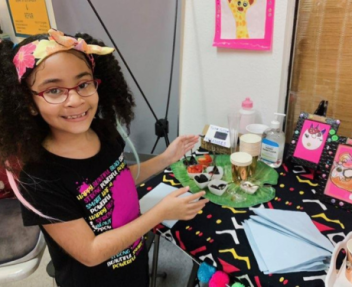 Nine year old girl with glasses and a floral headband smiles and points to her colorful artwork displayed on a table