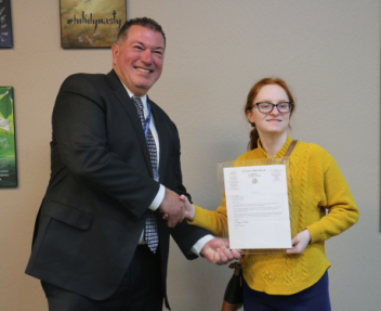 Superintendent Olmos stands next to Tori Berg in the Learning Center, holding the framed letter from Senator Niello in one hand and shaking Tori's hand with the other.