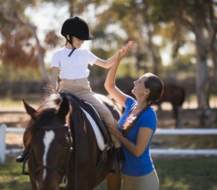 Side view of woman giving high five to girl sitting on horse in paddock, taking horseback riding lessons for enrichment activity