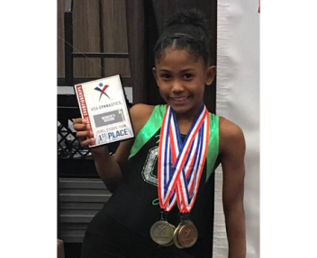 Female gymnast smiling at the camera holding 1st place award with gold medals