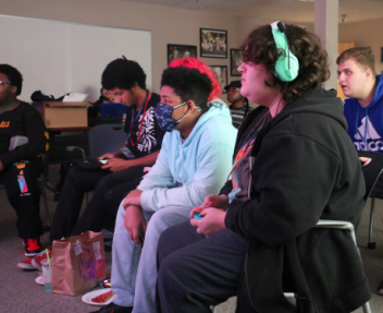 A group of students all sit around a projector screen and watch as one student plays the video game Super Smash Bros.