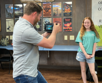 Camera man records smiling Visions student standing in front of a wall of published writing