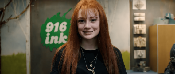 Smiling girl with red hair stands in front of a cheerful 916 ink sign, capturing a moment of happiness and excitement.